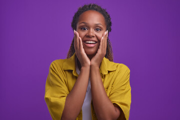 Wall Mural - Young positive curly African American woman millennial puts hands to cheeks and looks at camera with sincere smile hears romantic story or sees good deed stands on plain purple background