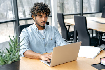 Serious focused confident indian or arabian man with curly hair, in elegant shirt, office manager, supervisor, sits at a desk in the modern office, works in a laptop, performs work tasks
