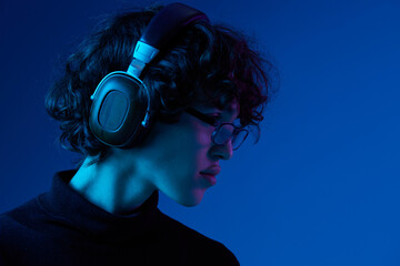 Wall Mural - Teenage man wearing headphones listening to music and dancing and singing open mouth with glasses, hipster lifestyle, portrait blue background, neon light, style and trends, mixed light, copy space