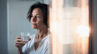 Mature age relaxed and content caucasian woman with short dark pixie hair drinking glass of water early in the morning in bathroom full of rising sun warm light rays. Healthy habit, morning routine