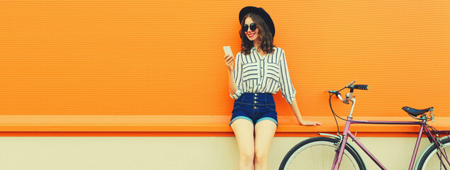 Summer image of happy smiling young woman with smartphone and bicycle on city street on orange background, blank copy space for advertising text