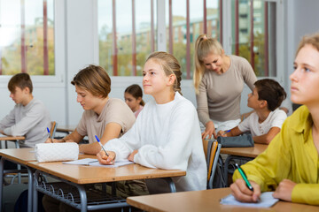 Wall Mural - Teenage boy and girl listening to teacher and writing exercises in notebook at lesson in secondary school