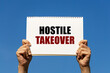 Hostile takeover text on notebook paper held by 2 hands with isolated blue sky background. This message can be used as business concept about hostile takeover.