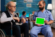 © DC Studio - Nursing home nurse holding horizontal green screen on digital tablet at appointment with elderly patient. Man in wheelchair in common room looking at device screen with copy space.