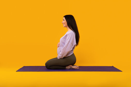 Wall Mural - Prenatal yoga. Pregnant lady sitting on fitness mat and touching belly, yellow studio background, side view