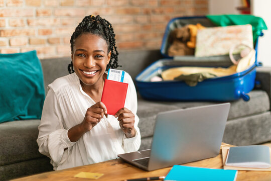 Wall Mural - Excited black lady planning vacation, sitting with passport and tickets in front of laptop and smiling at camera, ready for journey