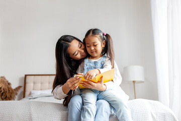 asian family sitting on bed and reading book at home, korean woman mother teaching little daughter to read book