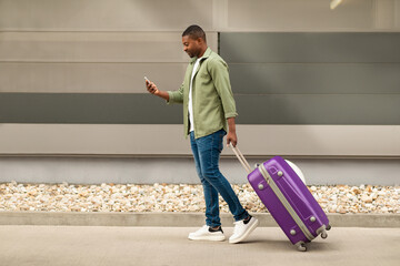 Wall Mural - Black Traveler Man Using Cellphone Walking With Travel Suitcase Outside