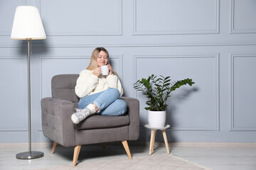 Poster - Young woman with cup of drink relaxing in armchair at home, space for text. Interior design