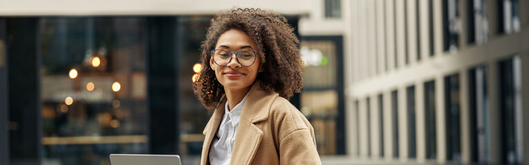 Happy african business woman working laptop while sitting outside on background of office building