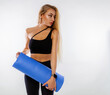 © oleg_ermak - Caucasian woman with long blond hair in black sportswear hold yoga mat or mat for fitness poses on a white background and shows perfect body. Personal care and sports at home.