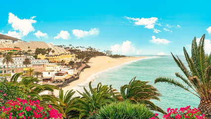 Wall Mural - Panoramic landscape of Morro Jable beach in Fuerteventura, Canary Islands, Spain, with palm trees and flowers in the foreground - Summer vacation background