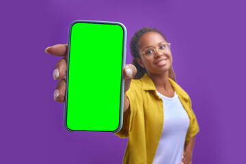 Young positive African American woman holds out green screen phone to camera to recommend cool mobile app or website for students stand on isolated purple background. Smartphone, technology