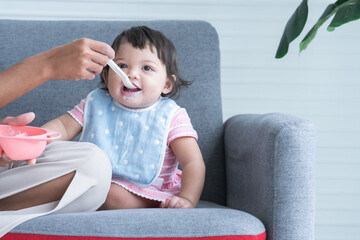 Caucasian Cute 7 month newborn baby girl eating blend mashed food, sitting on sofa with apron at home, mother hand is holding spoon feeding child, baby mouth mess up with food. Healhy food for kid