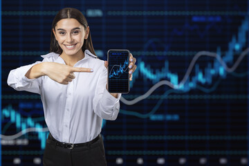 Wall Mural - Happy younng european businesswoman pointing at mobile phone with glowing candlestick forex chart and index on dark grid background. Trade, stock and finance concept. Double exposure.