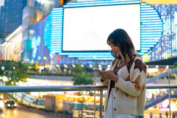 Wall Mural - Young Asian woman using smartphone for social media or online chat message in the city at night. Attractive girl enjoy and fun outdoor nightlife city street with using portable device technology.