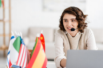 young language teacher in headset smiling during online lesson near laptop and international flags at home.