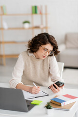 Wall Mural - curly student in glasses holding smartphone while taking notes near laptop at home.