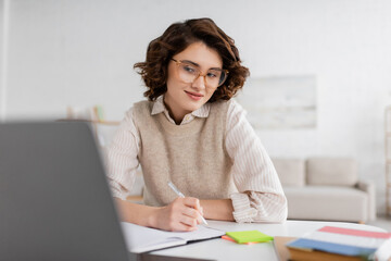 Wall Mural - smiling woman in glasses taking notes while learning foreign language and watching online lesson on 