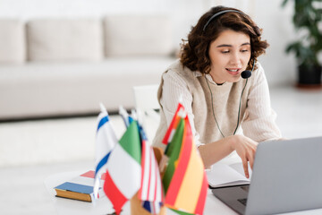 Wall Mural - positive language teacher in headset talking during online lesson on laptop near international flags