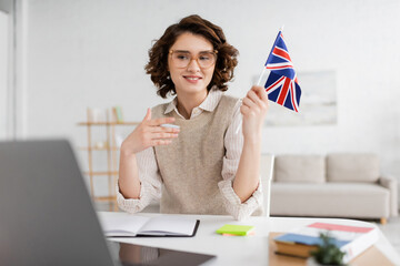 Wall Mural - young language teacher in eyeglasses holding flag of united kingdom near laptop on blurred foregroun