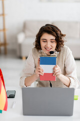 Wall Mural - joyful teacher in headset showing textbook of french language during online lesson on laptop at home