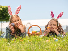 Little Girls Are Lying On The Green Grass Holding Painted Easter Eggs In Their Hands, Smiling, Looking Into The Frame, Egg Hunting Is A Traditional Game. Children's Fun