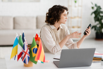 Wall Mural - young language teacher in glasses holding smartphone near laptop and international flags on blurred 