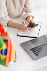 Wall Mural - cropped view of language teacher using smartphone near laptop and flags on blurred foreground.