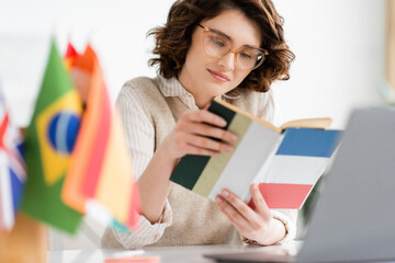 Wall Mural - pretty language teacher in glasses reading french dictionary book near international flags on blurre