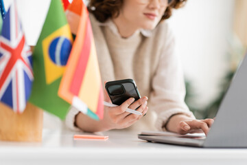 Wall Mural - cropped view of language teacher using mobile phone near laptop and different flags on blurred foreg
