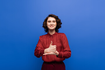 happy woman smiling at camera and showing help gesture on sign language isolated on blue.