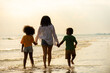 Happy African family mother and little daughter and son running and playing sea water together on the beach at summer sunset. Mom with child boy and girl enjoy outdoor lifestyle vacation at the sea