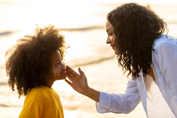 Happy African family on summer beach holiday vacation. Mother applying sunblock to little daughter. Mom and child girl kid enjoy outdoor lifestyle walking together on tropical beach at summer sunset.