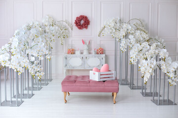 Wall Mural - Elegance white living room interior with pink sofa pouf and decorative blossom flowers orchids. Wedding decor. Easter festive decoration with flowers wreath on wall, ceramic rabbits and box with eggs.