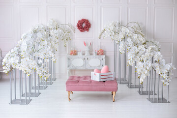 Wall Mural - Elegance white living room interior with pink sofa pouf and decorative blossom flowers orchids. Wedding decor. Easter festive decoration with flowers wreath on wall, ceramic rabbits and box with eggs.