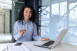 © Liubomir - Portrait of a young Muslim business woman sitting in the office at the table with a laptop. Holds a credit card, works with accounts, online shopping. She looks at the camera smiling.