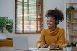 © SOMKID - Young African American businesswoman in wireless headset having web call using laptop sitting at home.