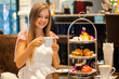 © TravelMedia - Woman in elegant dress enjoy English breakfast Afternoon tea in modern restaurant. Girl eating delicious sweet food. Cakes, sandwiches and macarons on high tea stand.