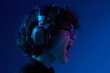 © SHOTPRIME STUDIO - Teenage man wearing headphones listening to music and dancing and singing open mouth with glasses, hipster lifestyle, portrait blue background, neon light, style and trends, mixed light, copy space