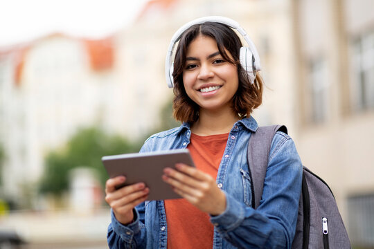 Wall Mural - Smiling Arab Female Student In Wireless Headphones Using Digital Tablet Outdoors