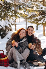  Happy young couple have a romantic picnic in the winter park. A young red-haired woman drinks tea and eats homemade cakes with her boyfriend in the middle of a snowy park.