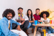 © unai - Group portrait of multiethnic friends eating popcorn and drinking soda on sofa. smiling looking at camera