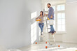 © Studio Romantic - Happy young couple painting the wall of their new home holding paint rollers and standing on the ladder. Married man and woman doing repair renovation preparing to move into a new flat.