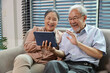 © feeling lucky - Happy smiling asian senior couple sitting on sofa and using tablet while online video call with friend or relative cousin at home living room. Internet information technology and lifestyle concept.