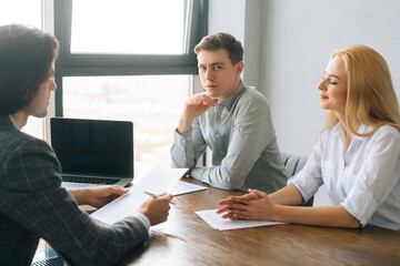 Side view of confident business partners discussing contract terms and project strategy at meeting in boardroom, sitting at table by window, three business people talking, sharing ideas at briefing.