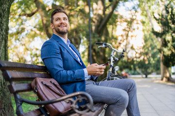 Handsome young businessman sitting on a bench in city park and using his mobile phone. bicycle is in blur.