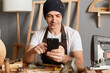 © sementsova321 - Indoor shot of handsome caucasian young adult man in cap and apron using his mobile phone in workshop, having break, chatting with friends, checking social networks.