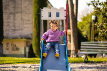 Sweet caucasian little toddler girl with two tails having fun smiling widely sliding down the yellow hill head first in the playground with violet sweeter sleeveless jacket jeans