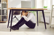 © Studio Romantic - Office worker hides under desk during earthquake with big magnitude. Scared worried fearful man sitting on floor under table holding head, hoping building won't collapse. Earthquake protection concept